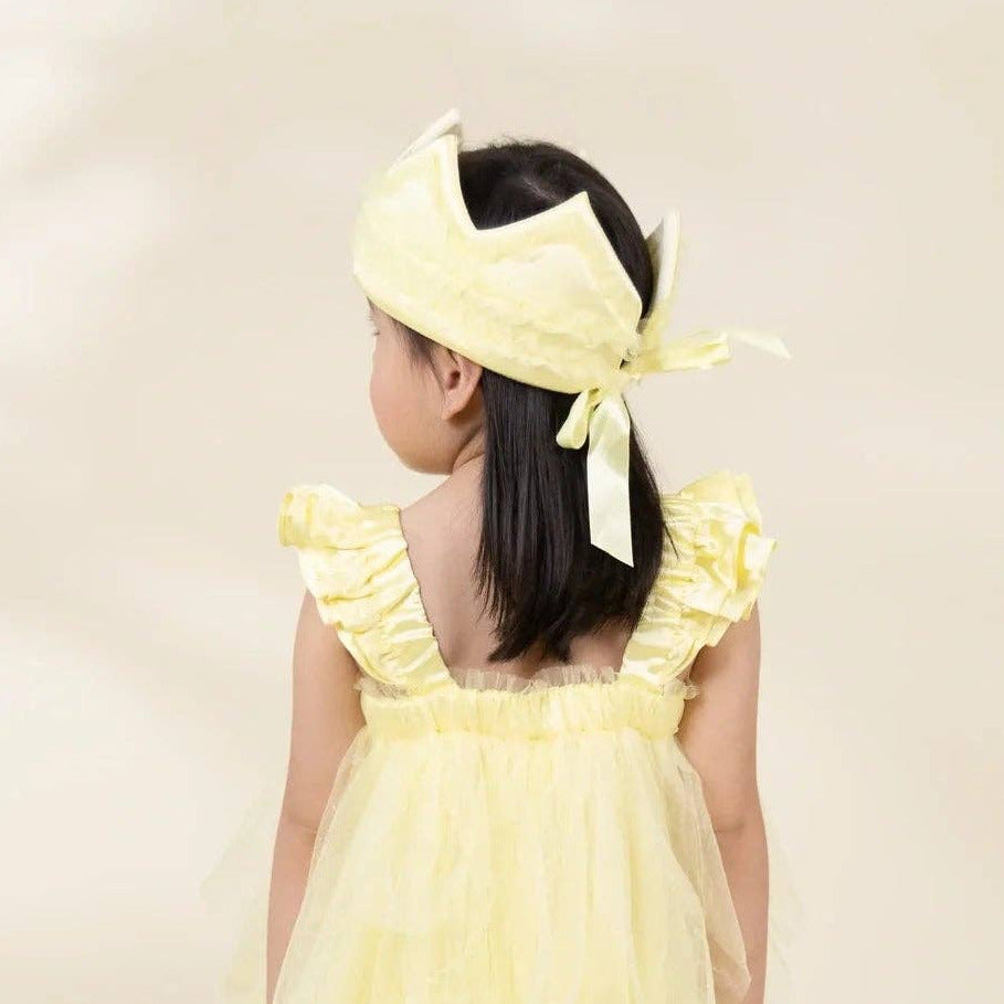 A young girl viewed from behind wearing a pale yellow ruffled dress with a layered tulle skirt and a matching yellow bow headband, standing against a soft cream studio background.