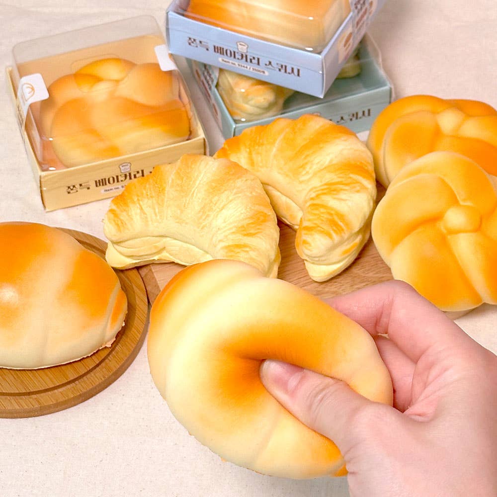 Hand holding a croissant-shaped bread with more bread items and packaging in the background.