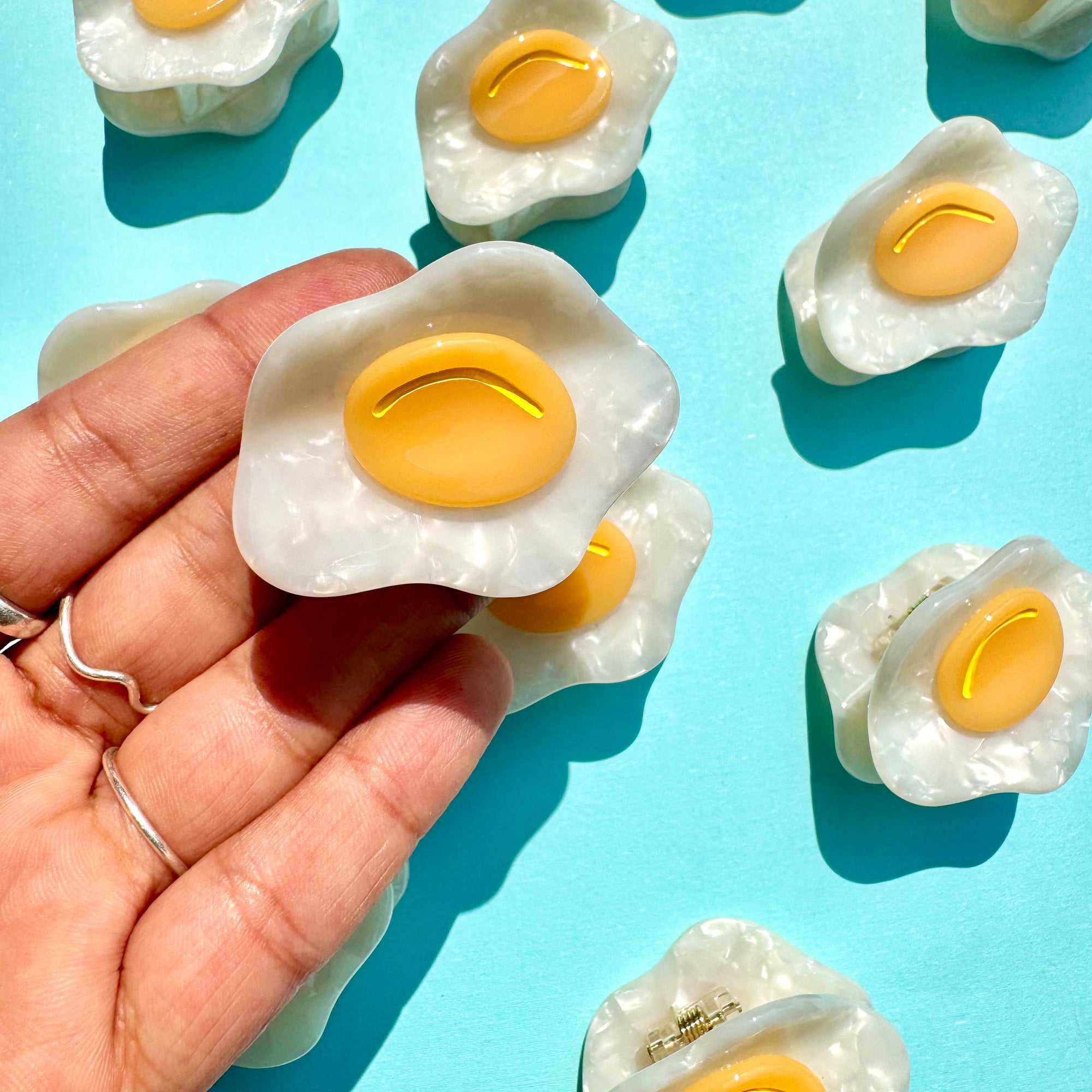 Hand holding a fried egg-shaped dessert against a blue background with more similar desserts.