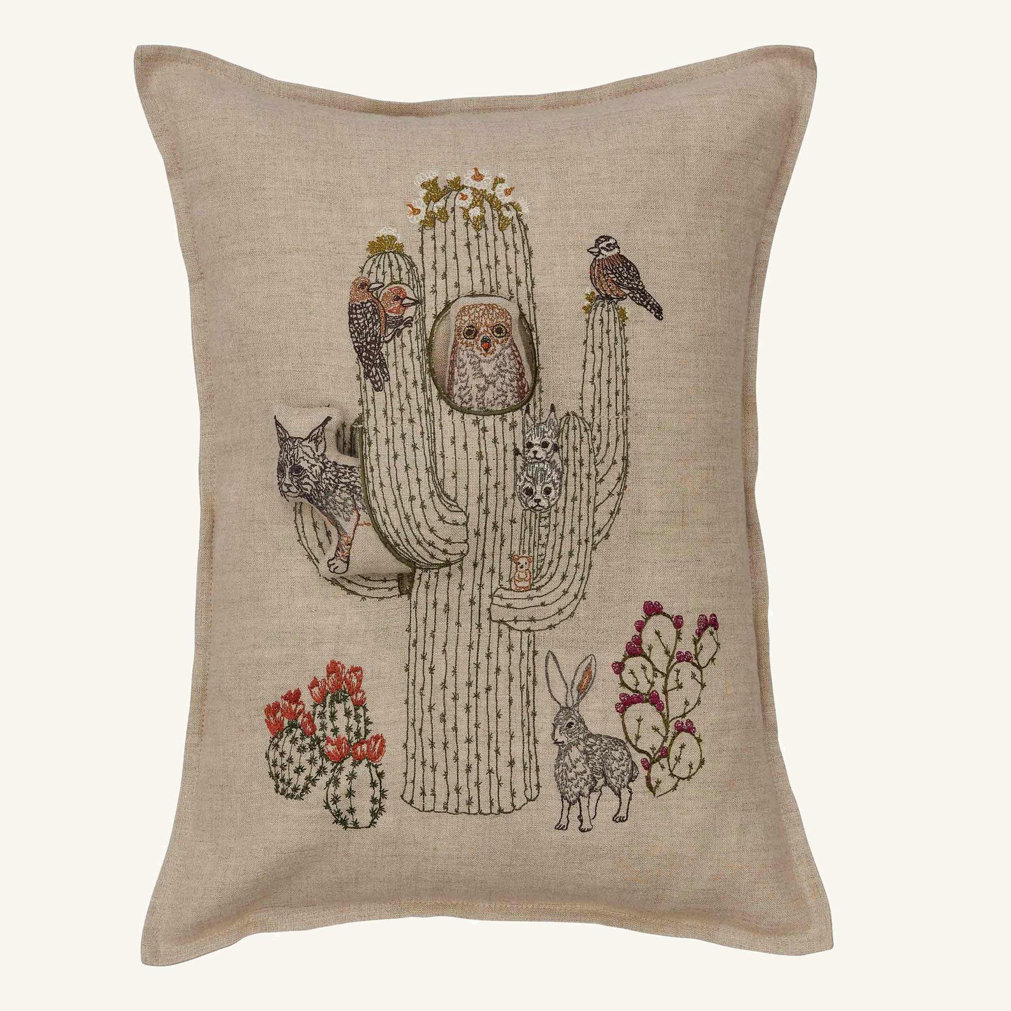 A square beige linen throw pillow featuring a detailed embroidered design of a saguaro cactus with owls, birds, a rabbit, and prickly pear cacti in muted earth tones.