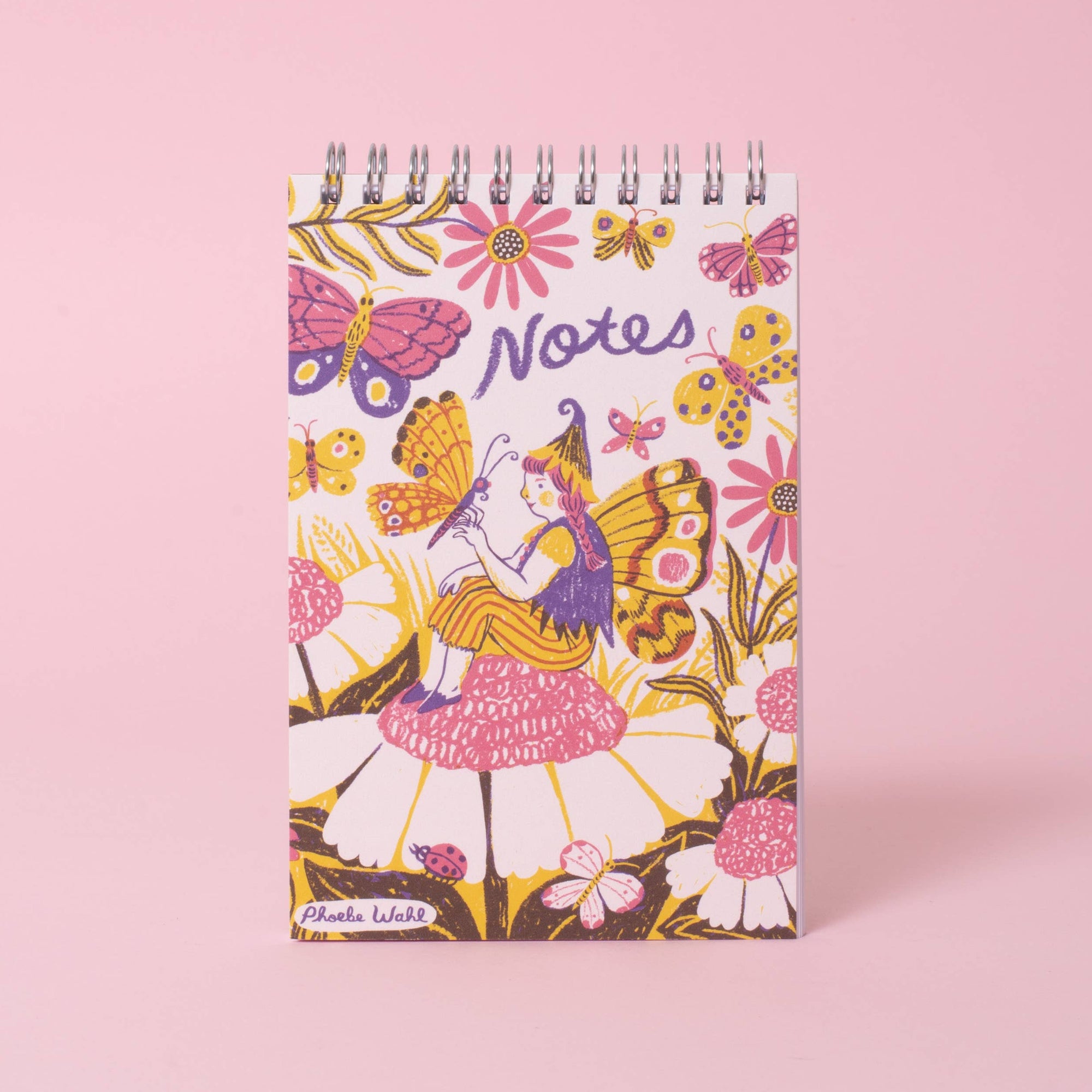 A spiral-bound notebook with a whimsical cover illustration featuring butterflies, flowers, and a fairy, standing upright on a solid pink background.