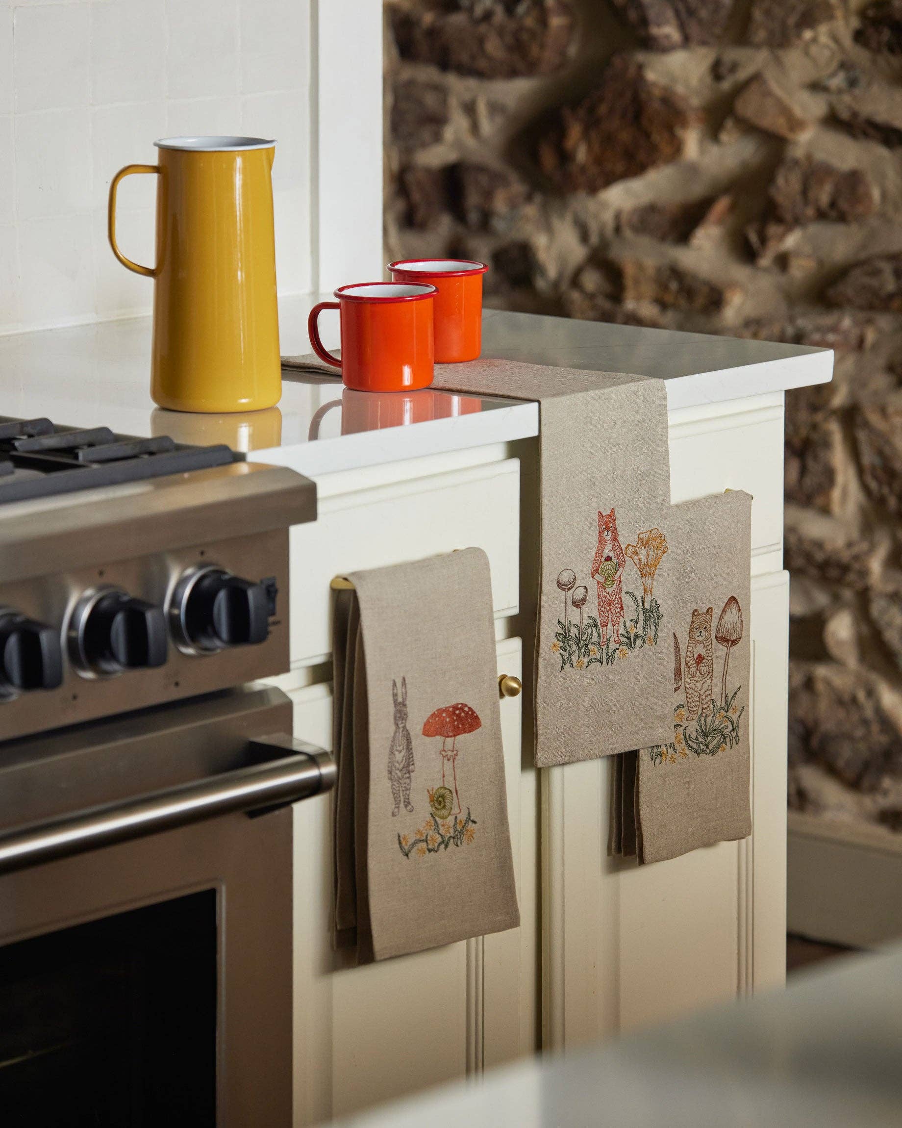 Kitchen scene with a stove, mugs, and decorative towels on a countertop.