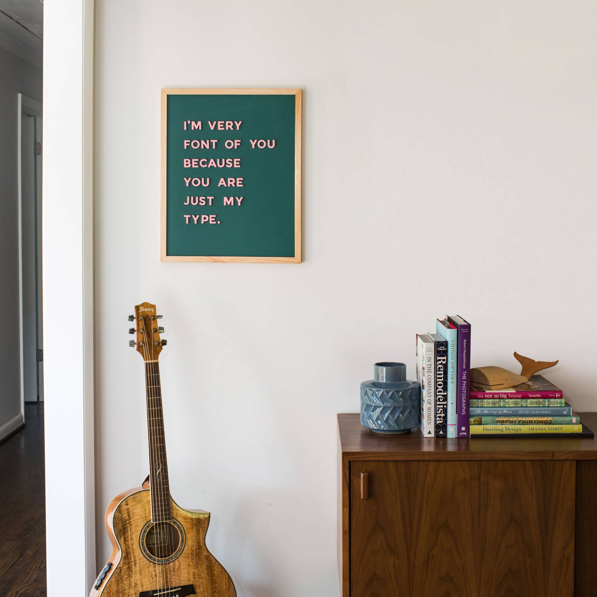 Framed quote on a wall above a guitar and books on a shelf.