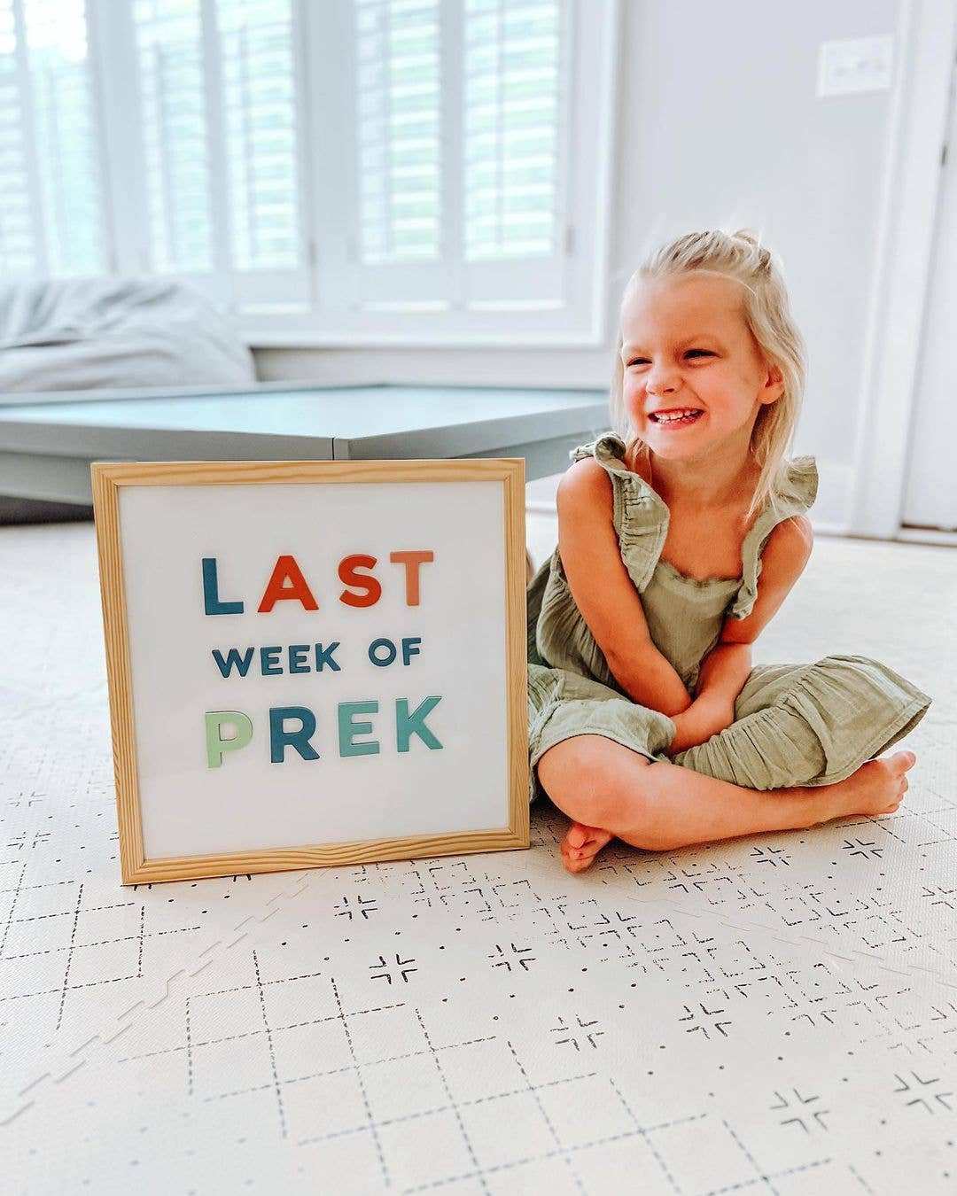 Child sitting on a floor with a sign reading 'Last Week of PreK' in a bright room.