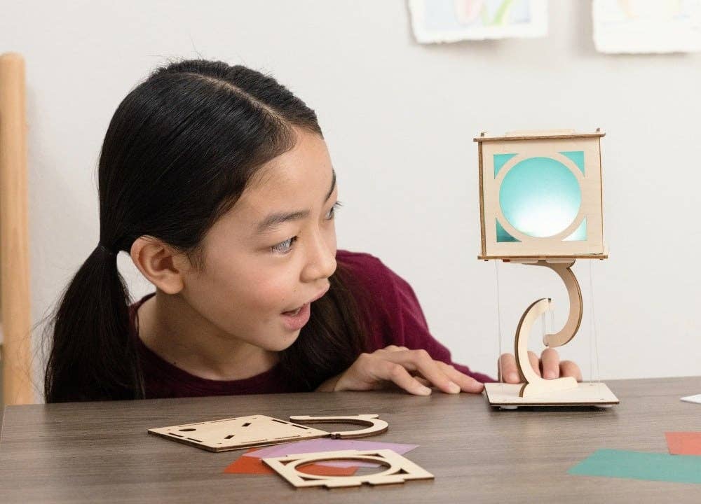 Young girl interacting with a wooden lamp and geometric shapes on a table.