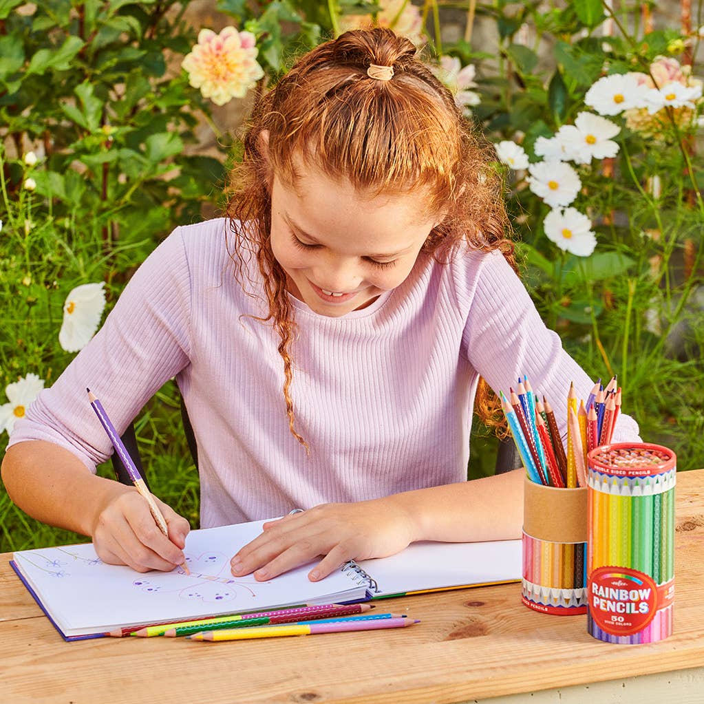 Young girl drawing with colored pencils outdoors on a wooden table with flowers in the background