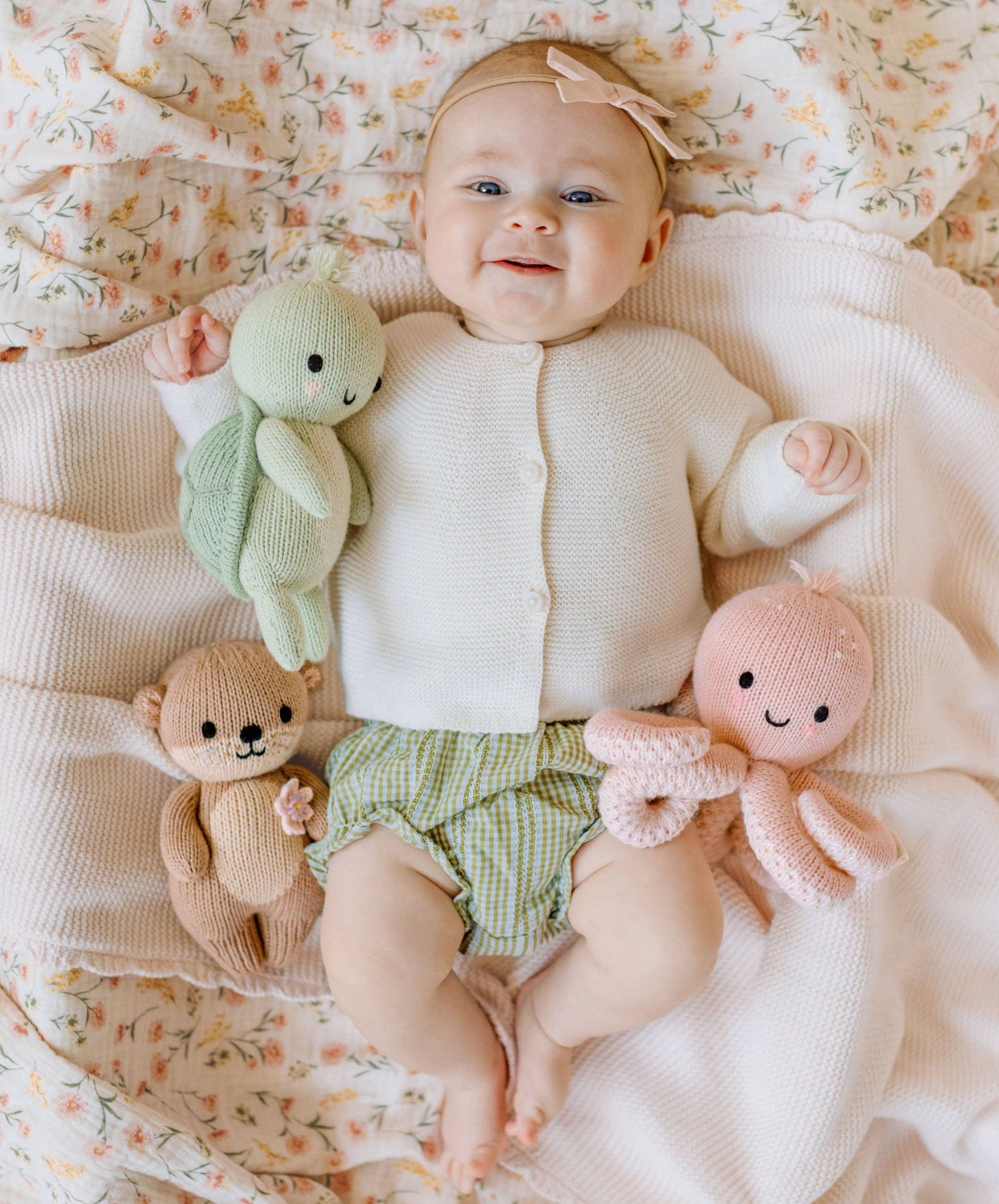 Baby lying on a floral blanket with three stuffed toys, wearing a white sweater and green checkered shorts.