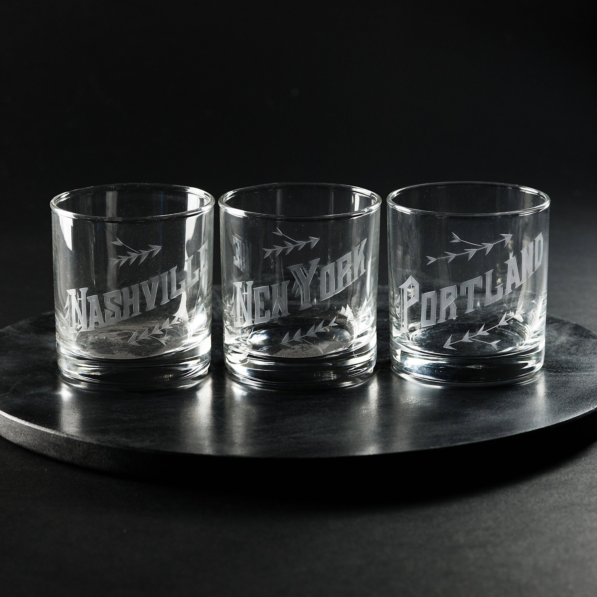Three glass tumblers with engraved city names on a dark surface