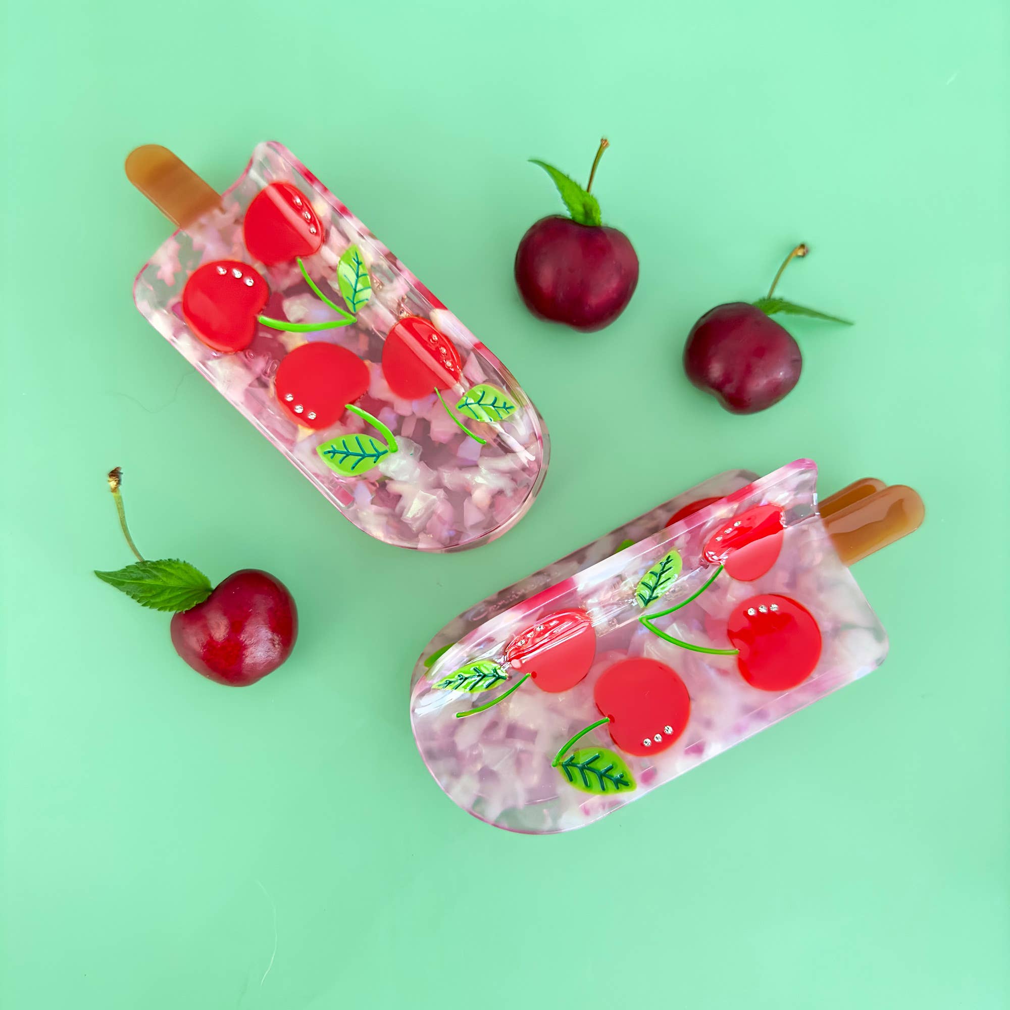 Two cherry-themed ice cream bars with red cherries and green leaves on a light green background.