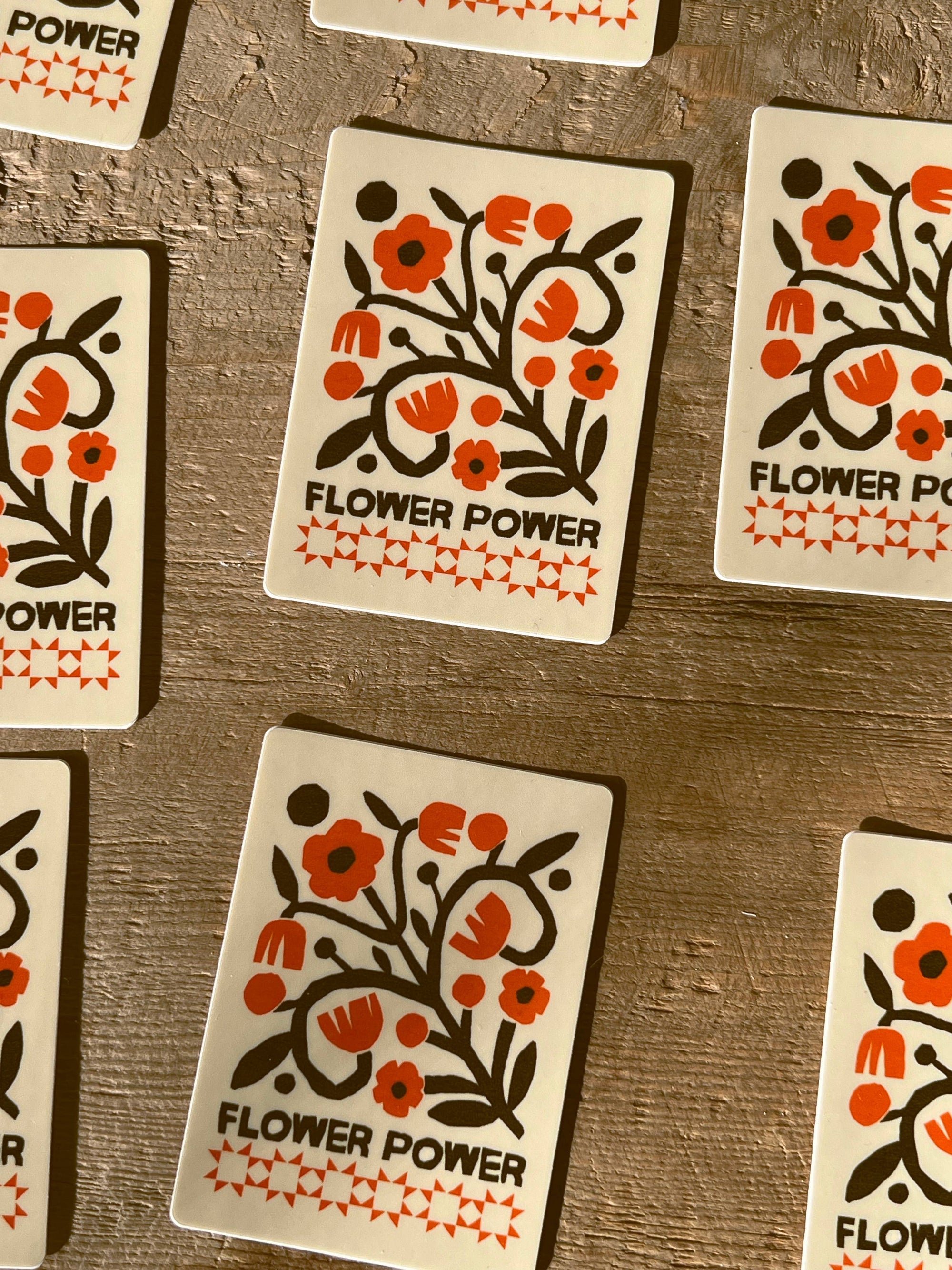 A close-up overhead view of several square cards laid on a rustic wooden surface. Each card has a cream background with a stylized floral illustration in orange and black and the words “FLOWER POWER” printed near the bottom.