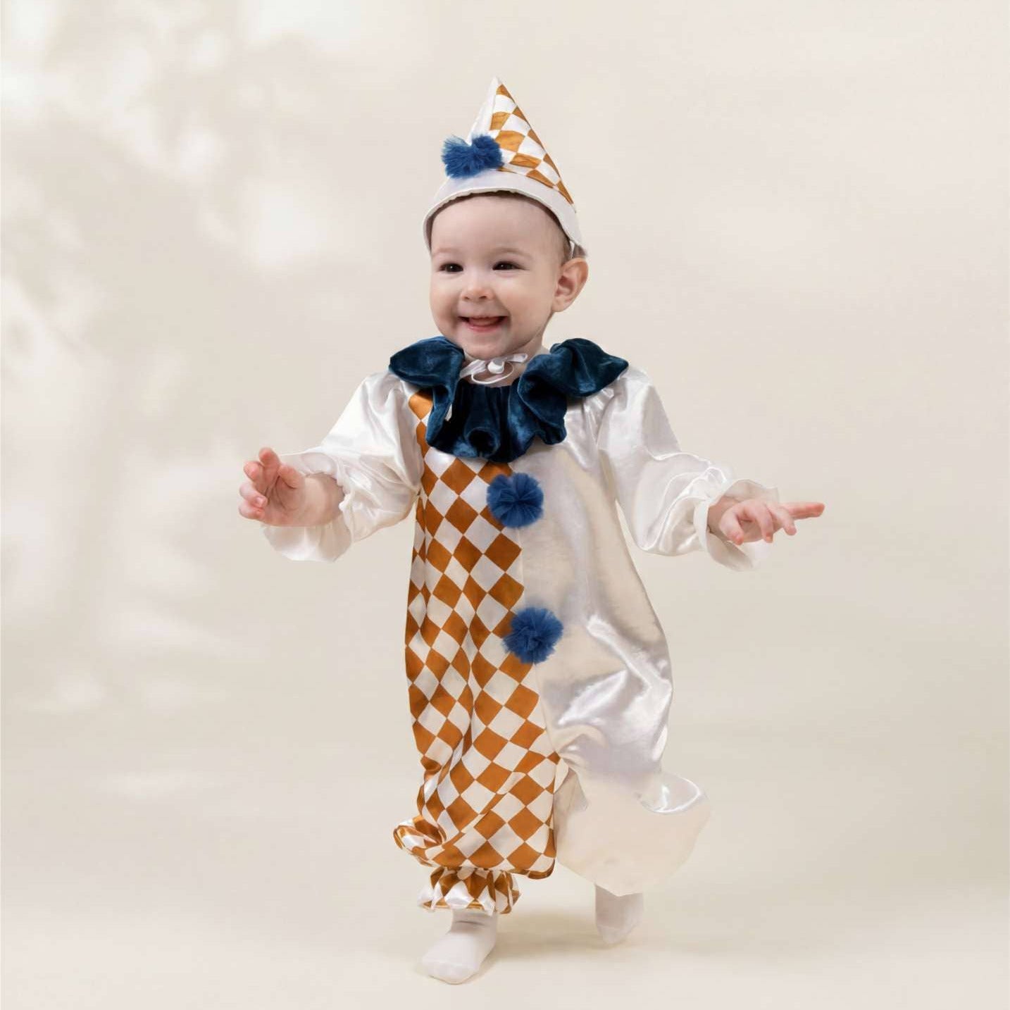 A baby wearing a white and gold checkered clown costume with a navy blue bow tie and matching pointed hat, smiling and posing against a soft beige background.
