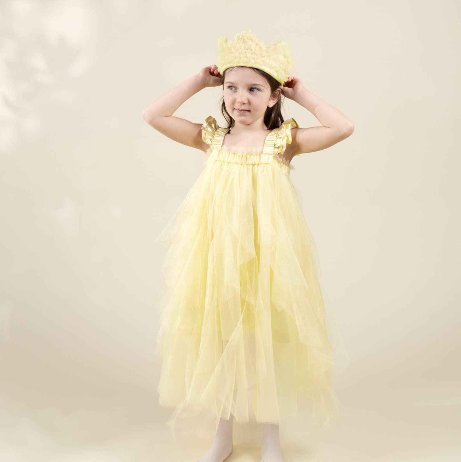A young girl modeling a light yellow tulle princess dress with gold trim and a matching yellow crown, standing against a soft beige studio backdrop.