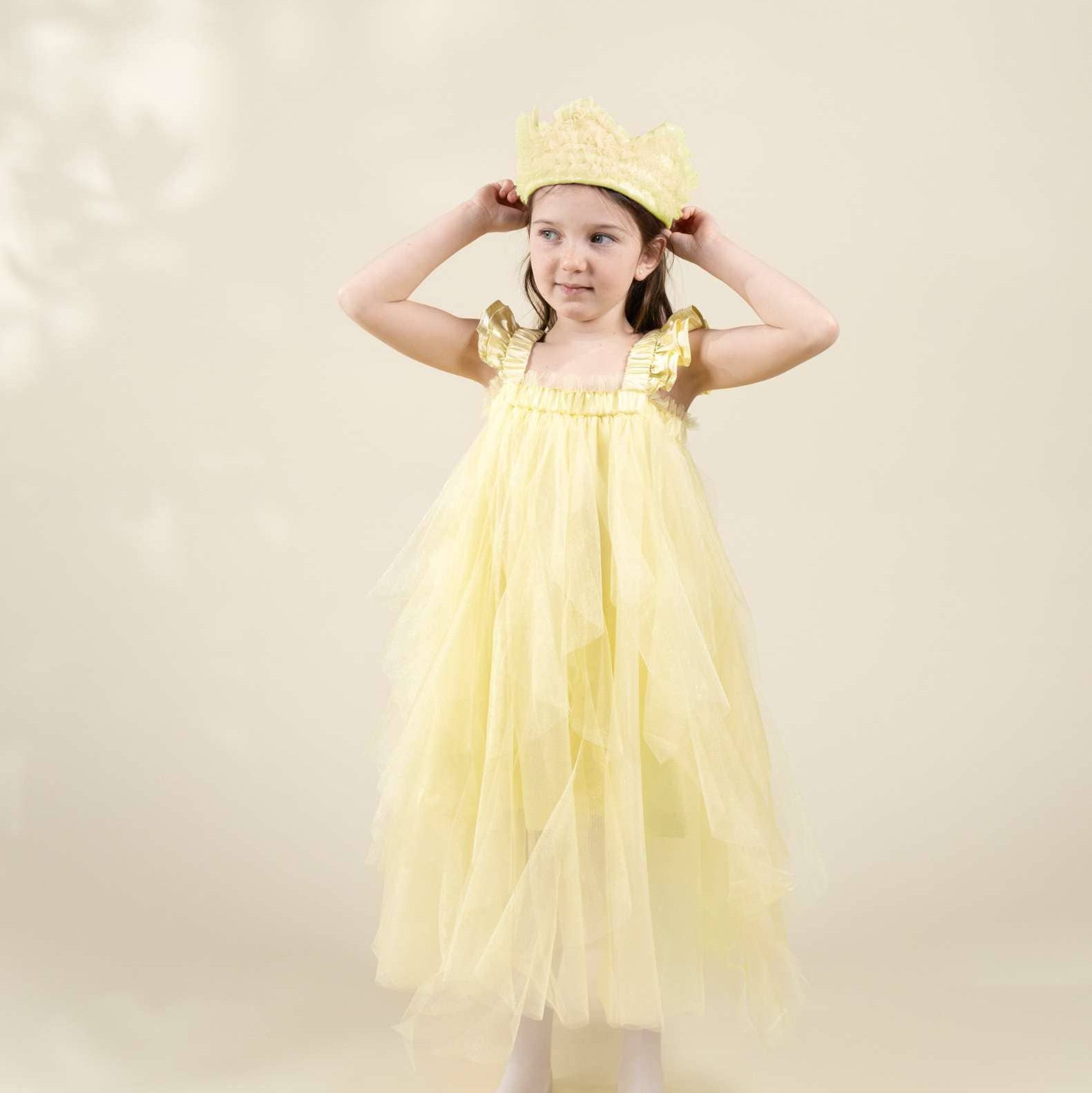 A young girl modeling a light yellow tulle princess dress with gold trim and a matching yellow crown, standing against a soft beige studio backdrop.