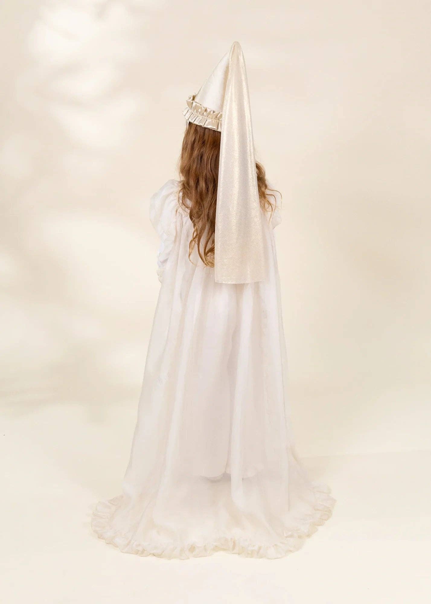 A child viewed from behind wearing a white or ivory pointed habit with a long flowing train, a matching veil, and a jeweled crown against a soft cream studio background.