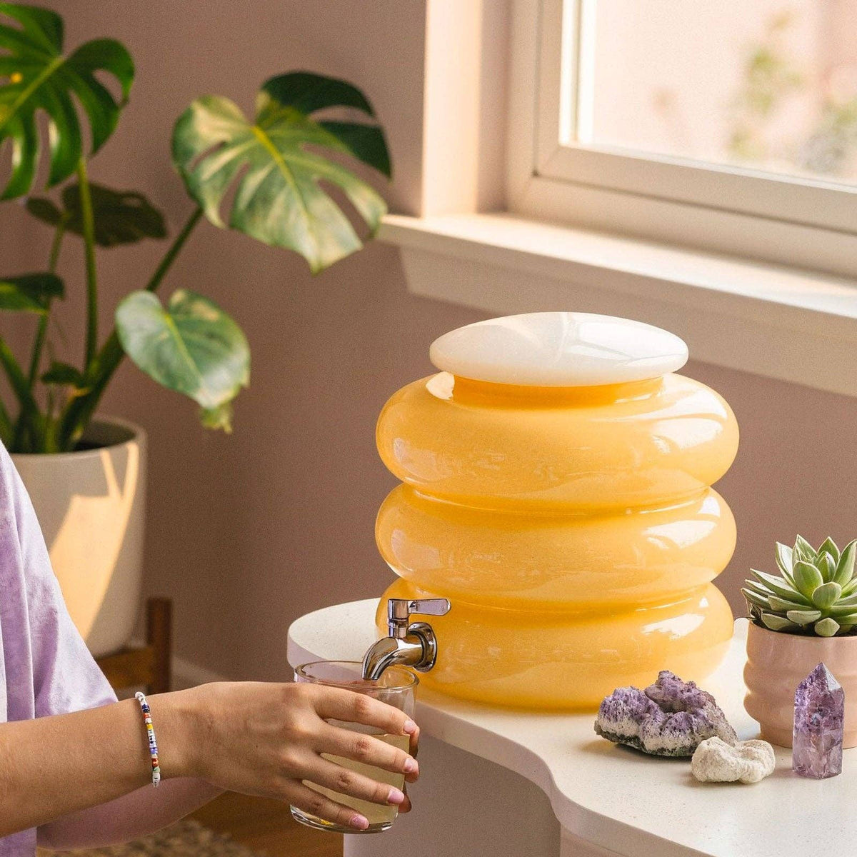 Person using a yellow water dispenser on a table with plants and decor in the background