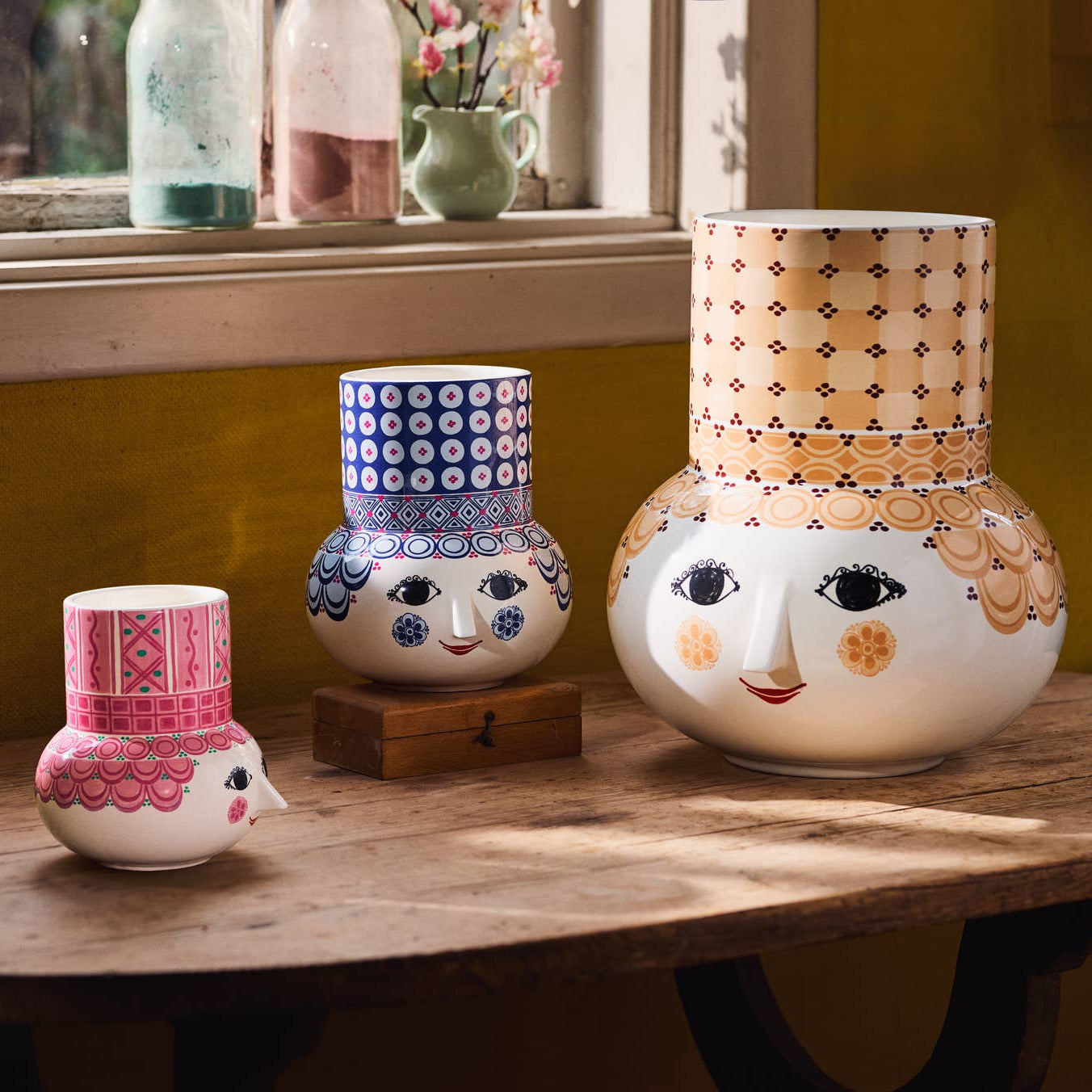 Three decorative vases with face designs on a wooden table near a window.