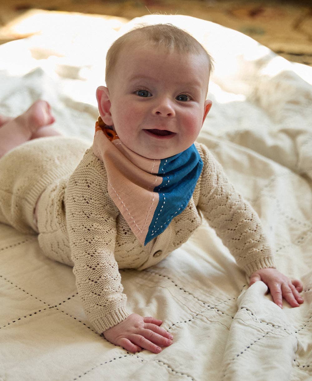 Baby wearing a beige knitted outfit with a blue bandana on a soft surface