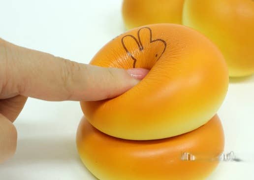 A close-up of a finger pressing down on a soft, squishy orange bagel-shaped toy or stress relief item, with another similar item stacked below and more blurred items in the background.