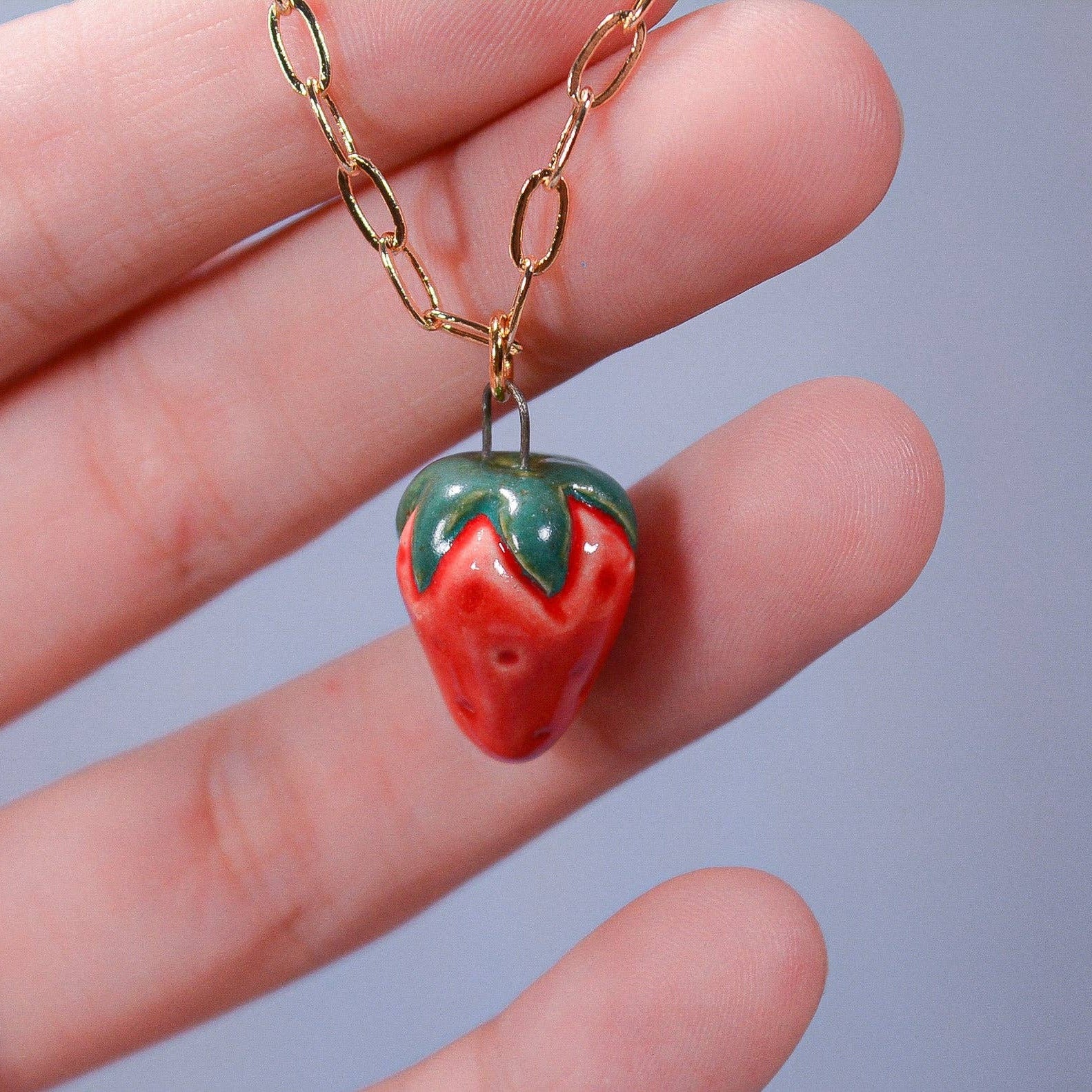 Strawberry-shaped pendant on a gold chain held in a hand against a gray background