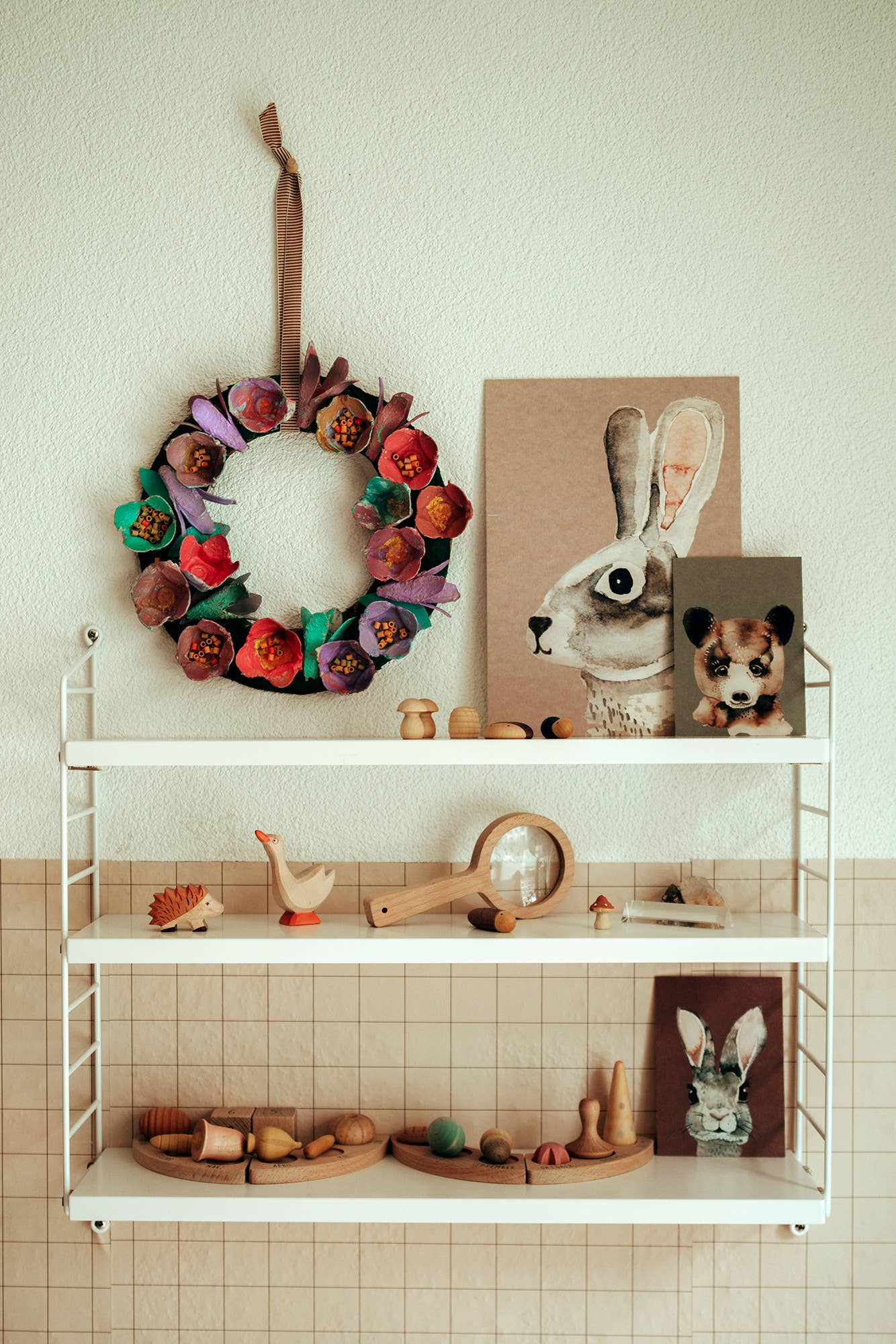 A styled nursery or playroom shelf display featuring a handmade fabric flower wreath hanging on a wall, alongside a rabbit portrait print, a stuffed animal, and wooden toy accessories on white wire shelving.
