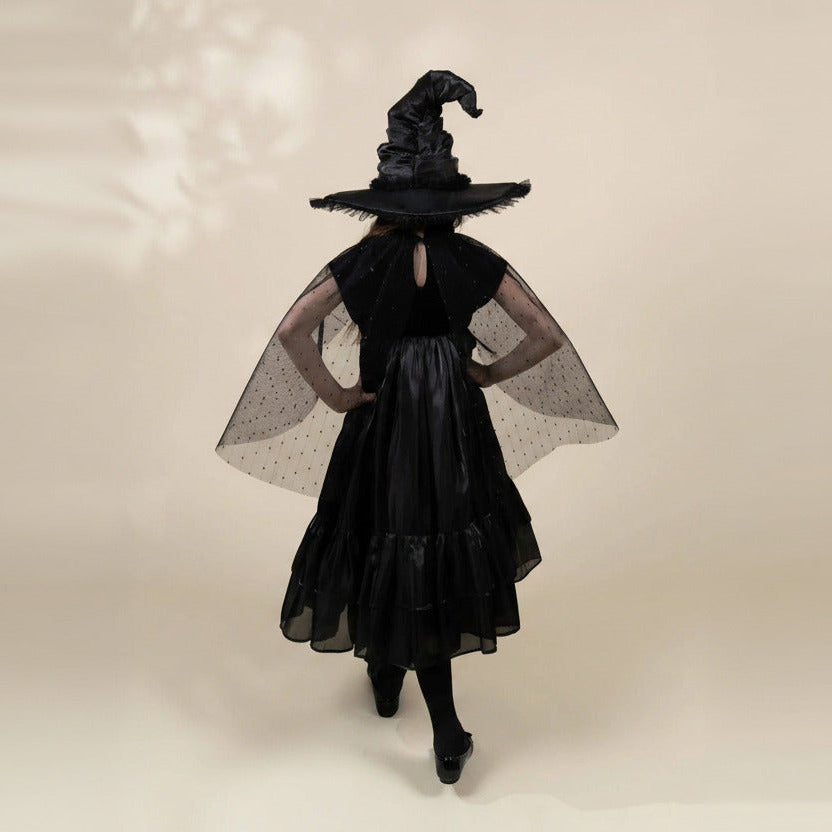 A model from behind wearing a black witch costume, featuring a tall pointed hat, a tiered black dress, sheer black polka-dot cape wings, and black tights, set against a plain beige background.