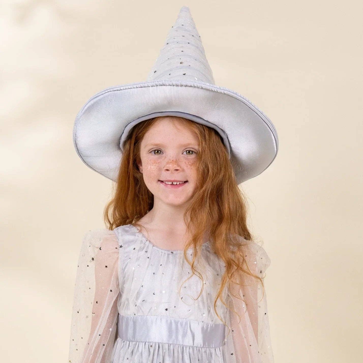A young girl with long red hair wearing a white witch costume dress with sheer sleeves and a matching pointed witch hat, both decorated with small black polka dots, set against a solid beige background.
