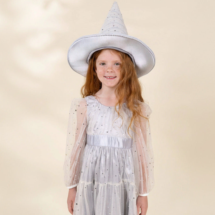 A young girl with long red hair wearing a white witch costume dress with sheer sleeves and a matching pointed witch hat, both decorated with small black polka dots, set against a solid beige background.