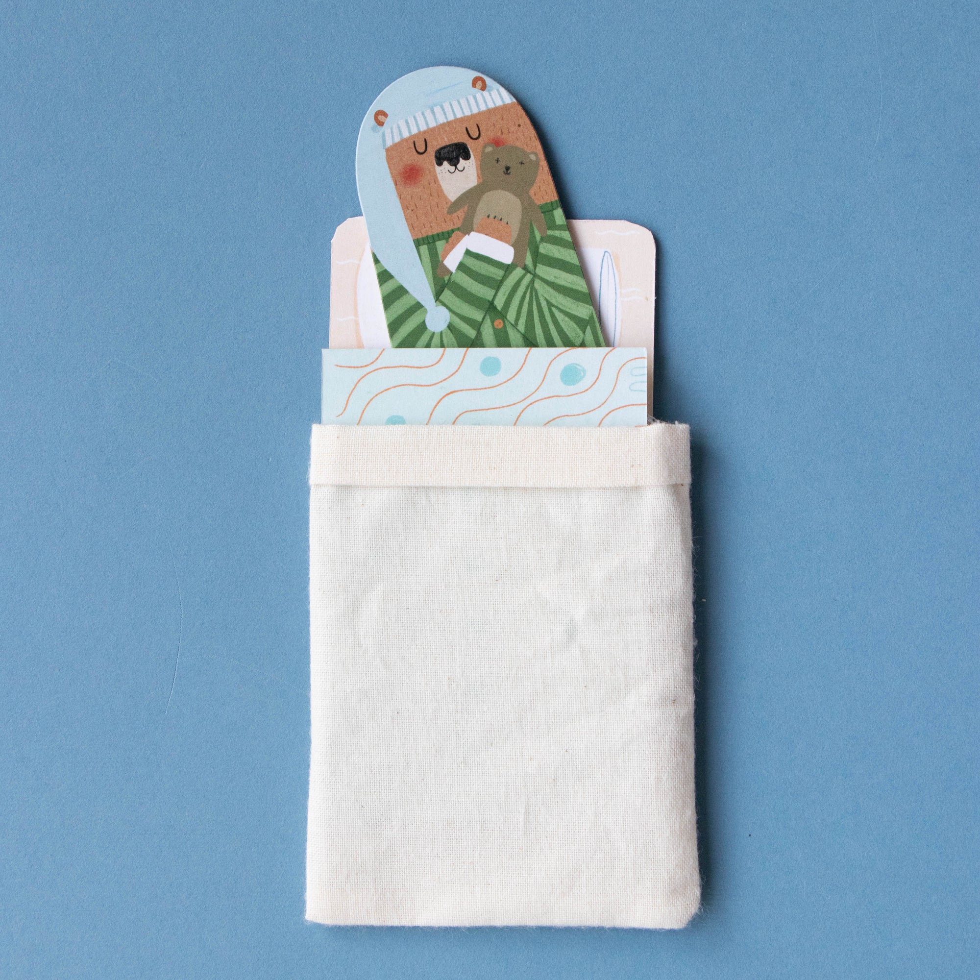 Cardboard bear toy with a green sweater peeking out of a white pocket against a blue background