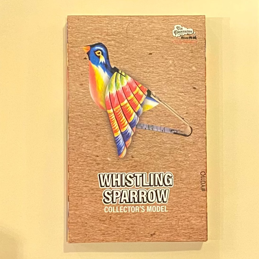 Colorful bird model on a cardboard backing with 'Whistling Sparrow' text on a wooden surface