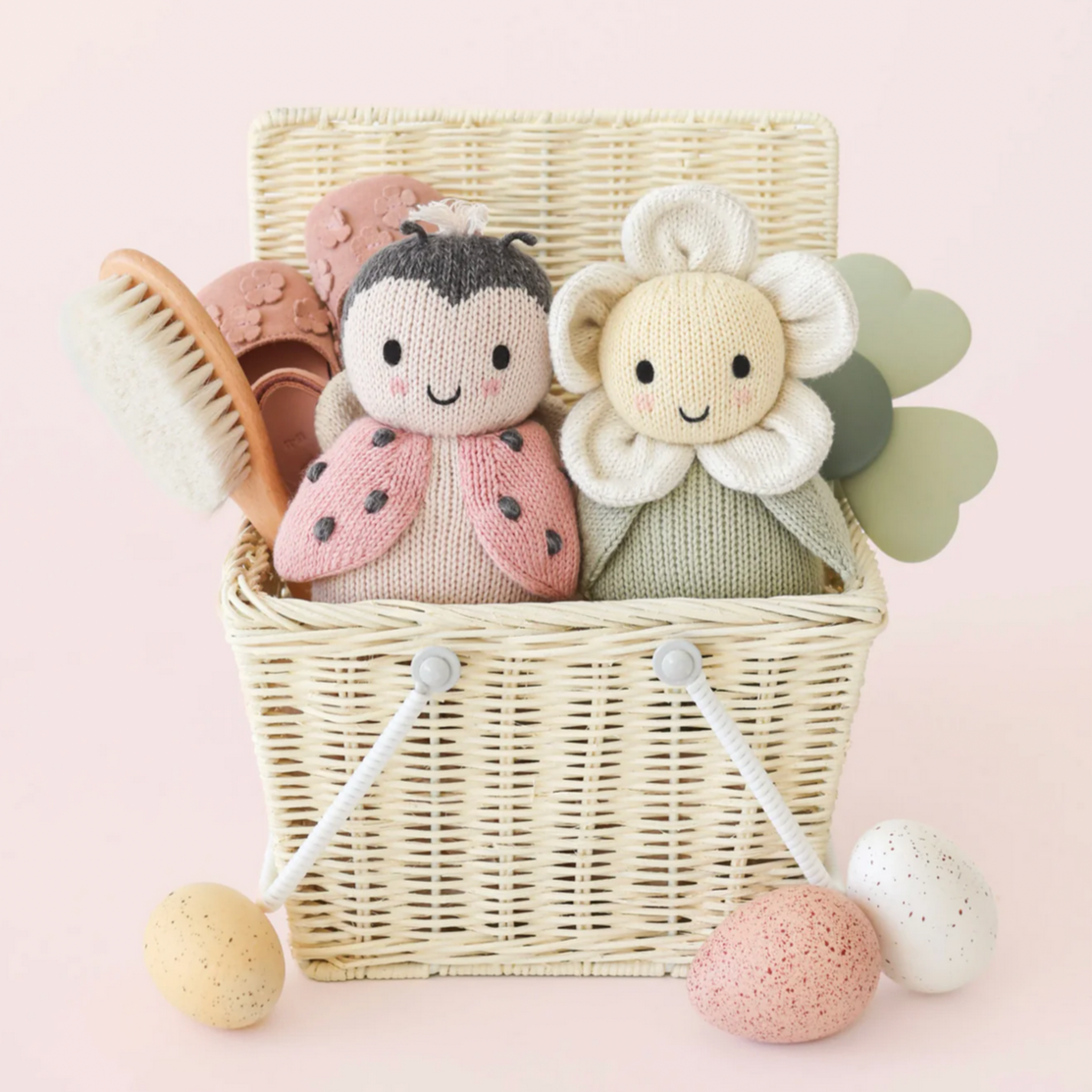 Two soft toys, one resembling a flower and the other a ladybug, in a wicker basket on a pink background.