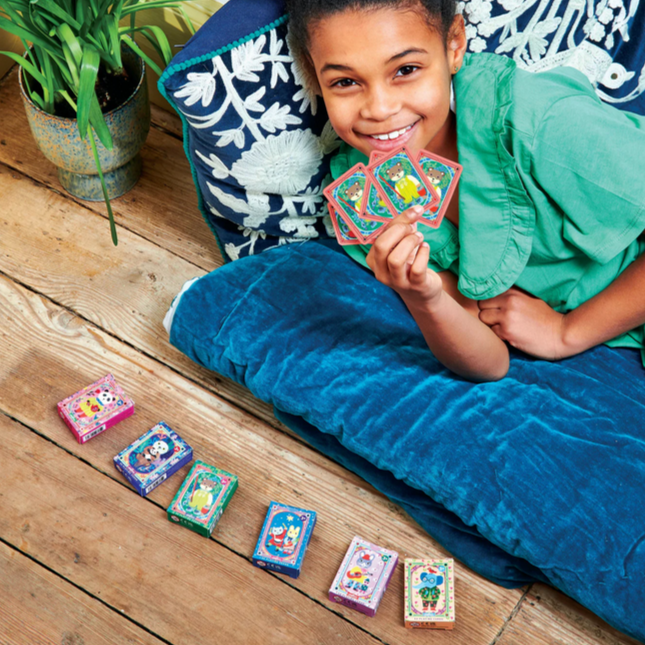 Child sitting on a blue cushion holding colorful cards on a wooden floor with plants in the background.