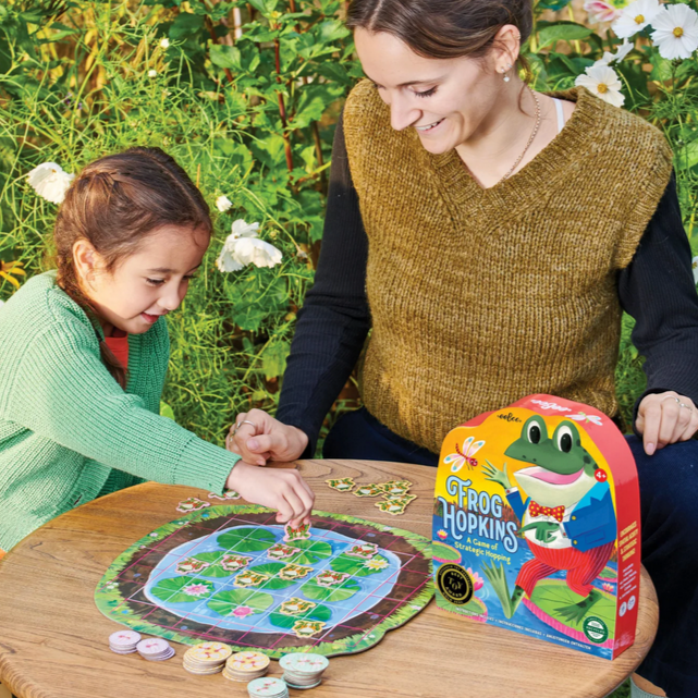 Woman and child playing a board game outdoors with a 'Hoppy Hopkins' game box.