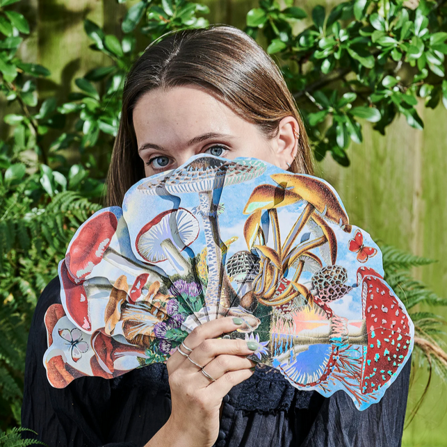 Woman holding a colorful fan with nature-themed design against a green leafy background