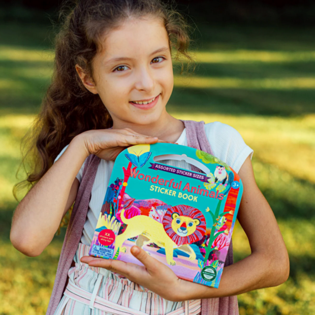 Young girl holding a colorful sticker book with animals on a grassy background