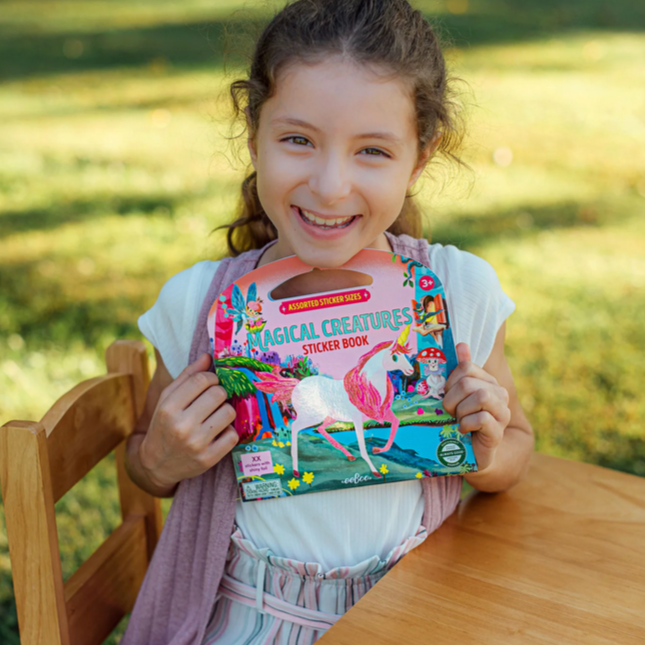 Young girl holding a sticker book with a colorful cover, sitting outdoors.