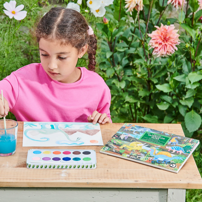 Child at a table with art supplies outdoors, surrounded by nature.