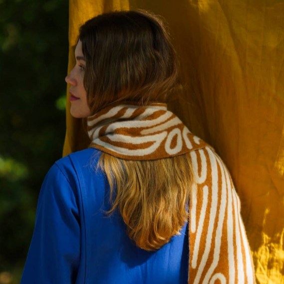 Person wearing a blue coat and patterned scarf standing against a yellow curtain.