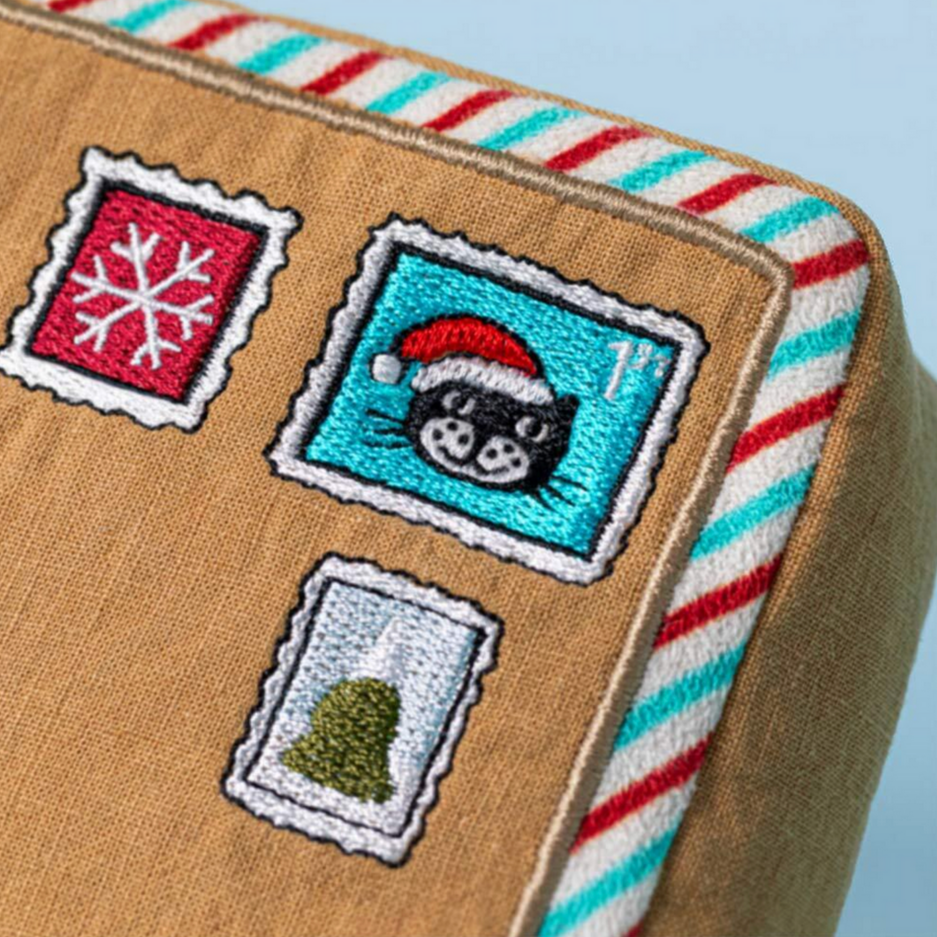 Close-up of a gingerbread house-shaped pillow with embroidered patches on a light blue background.