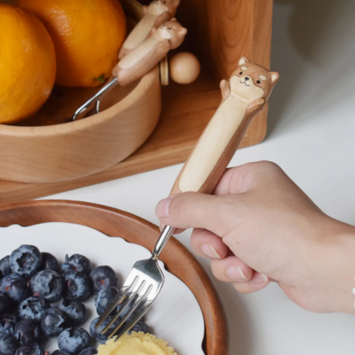 Hand holding a dog-shaped fork over a plate of blueberries with a wooden bowl in the background.