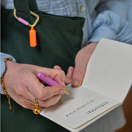 Person writing in a notebook with a pink pen that's clipped to a green bag 