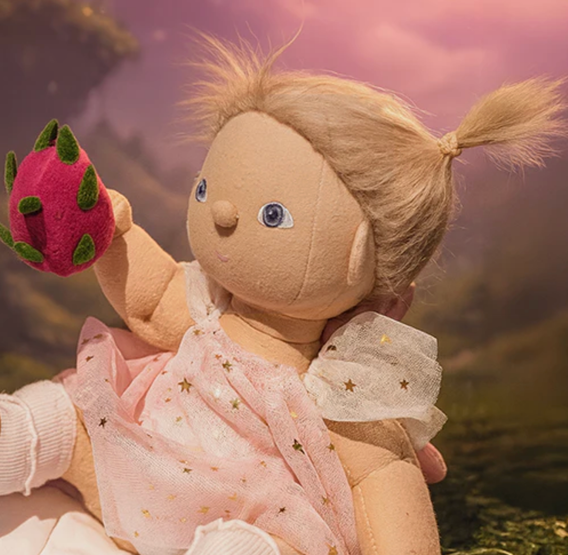 Stuffed doll with blonde hair and blue eyes holding a pink flower against a blurred natural background