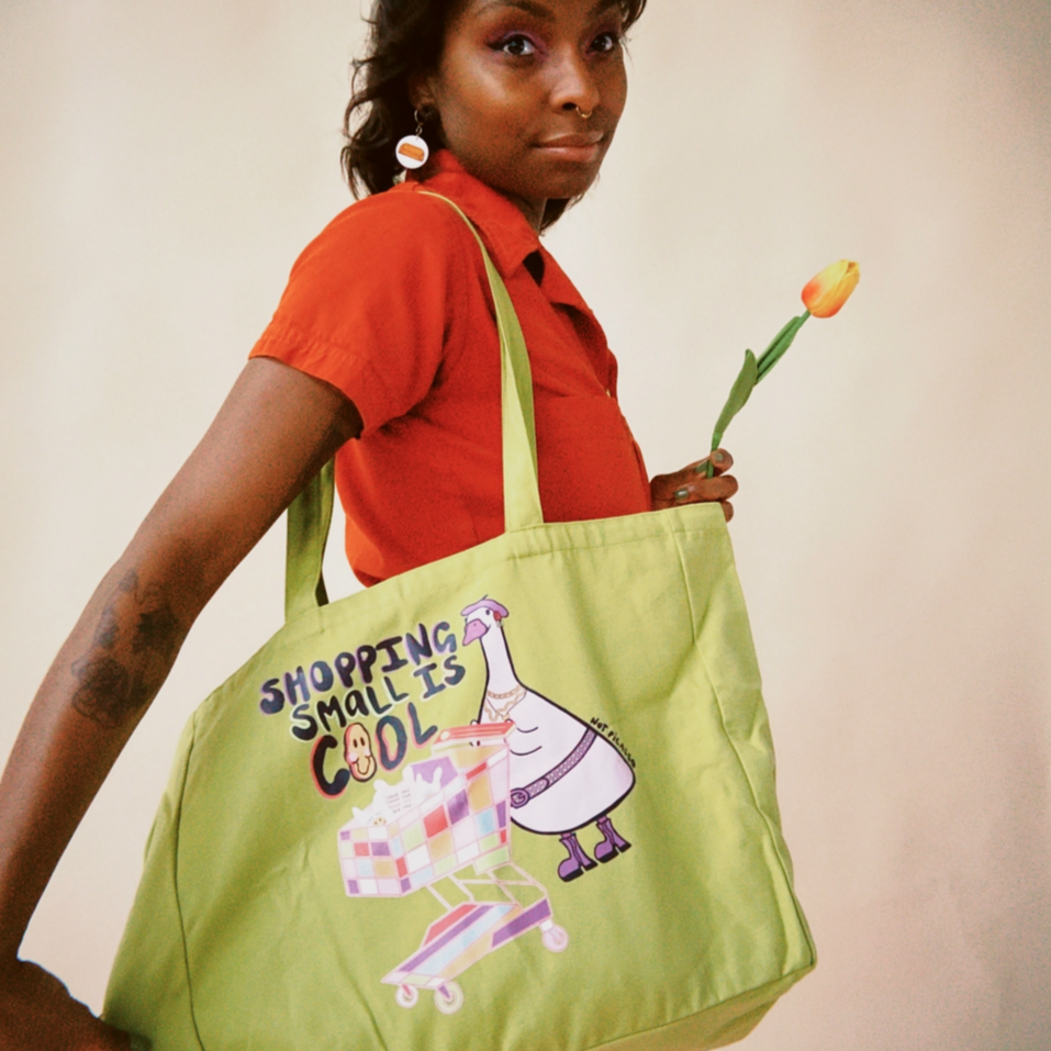 Person holding a green tote bag with a cartoon character and text, wearing an orange shirt and red pants.