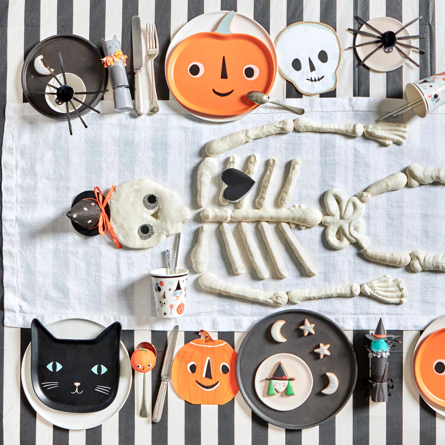 Halloween-themed table setting with decorative plates, cutlery, and skeleton figurines on a striped tablecloth.