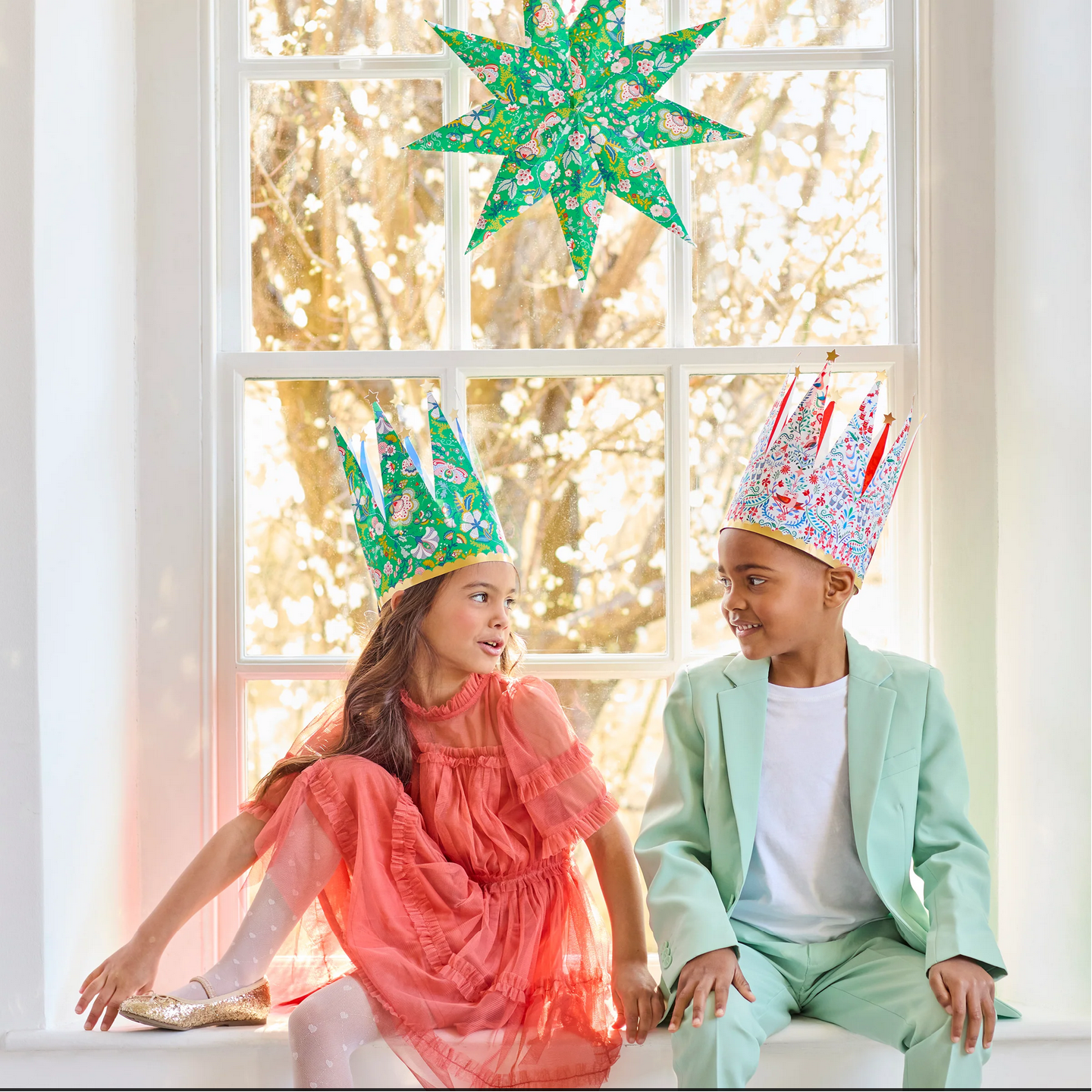 Two children wearing festive crowns sitting in front of a window with decorative lights.