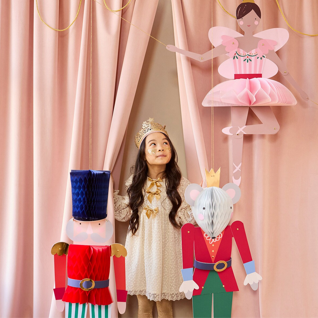 Child standing behind decorative cutouts of a ballerina, nutcracker, and mouse king with pink curtains in the background.