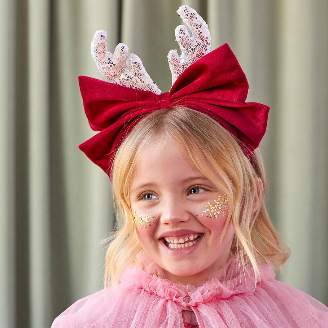 Young girl wearing a red bow headband with decorative antlers, smiling against a neutral background.