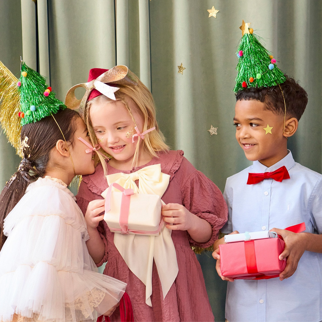 Three children wearing festive hats and holding Christmas presents against a curtain background.
