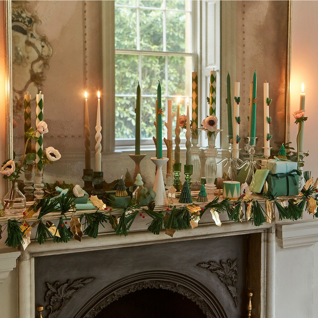 Decorative mantel with candles, flowers, and greenery in a room with large windows.