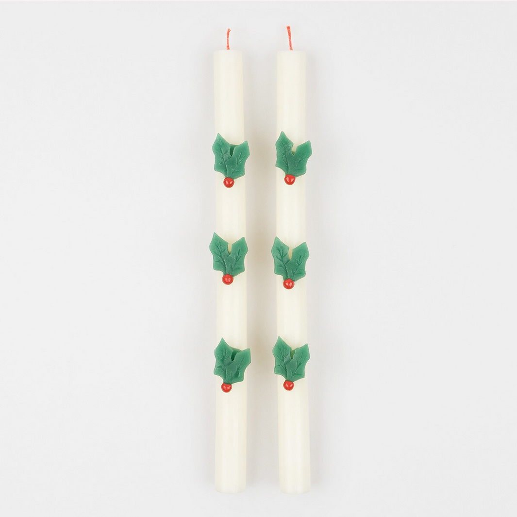 Two white candles with holly berry decorations on a light gray background