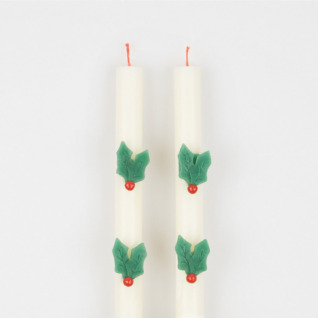 Two white candles with green leaf and red berry decorations on a light gray background